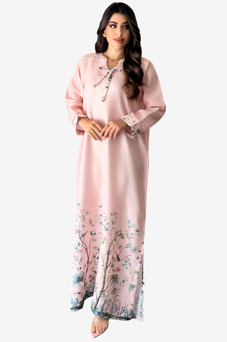 A woman modeling a long light pink kaftan dress with a floral print concentrated along the hem and small embroidered details near the neckline and cuffs, set against a plain light gray background.