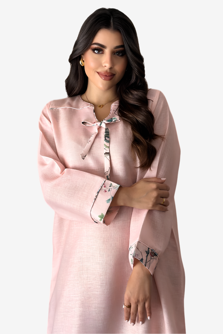 A woman modeling a light pink long-sleeve tunic or kurta with a floral print tie-neck bow and matching floral cuff accents, set against a plain light gray studio background.