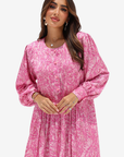 A woman modeling a pink long-sleeve dress with a white abstract floral print, featuring a buttoned neckline and a gathered, flowing skirt, set against a plain light gray background.