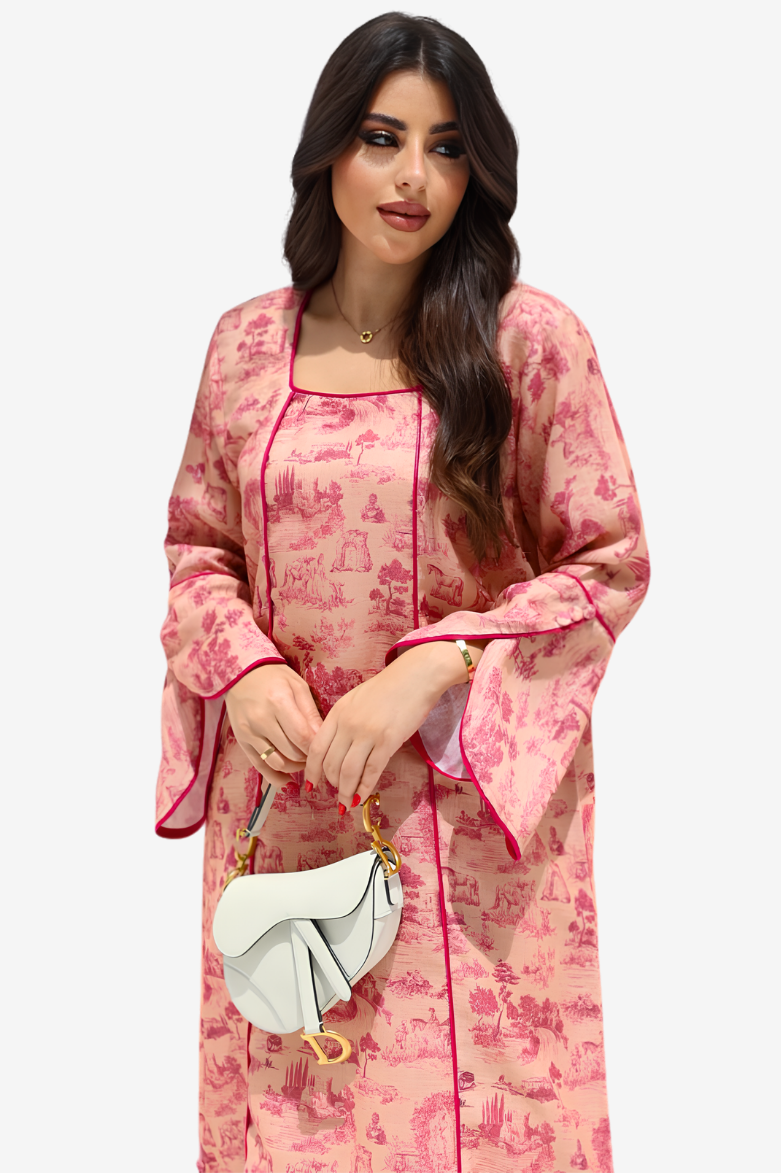 A woman modeling a pink and red toile-print kaftan dress with bright pink piping, holding a white handbag against a light gray background.