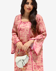 A woman modeling a pink and red toile-print kaftan dress with bright pink piping, holding a white handbag against a light gray background.