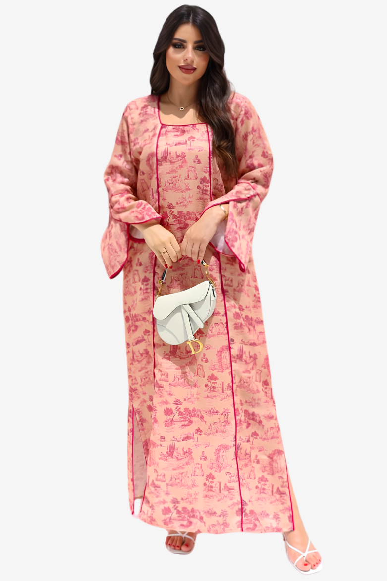 A woman modeling a long pink kaftan dress with a darker pink floral toile pattern, holding a small white handbag against a light gray background.