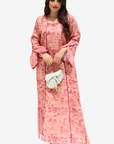 A woman modeling a long pink kaftan dress with a darker pink floral toile pattern, holding a small white handbag against a light gray background.