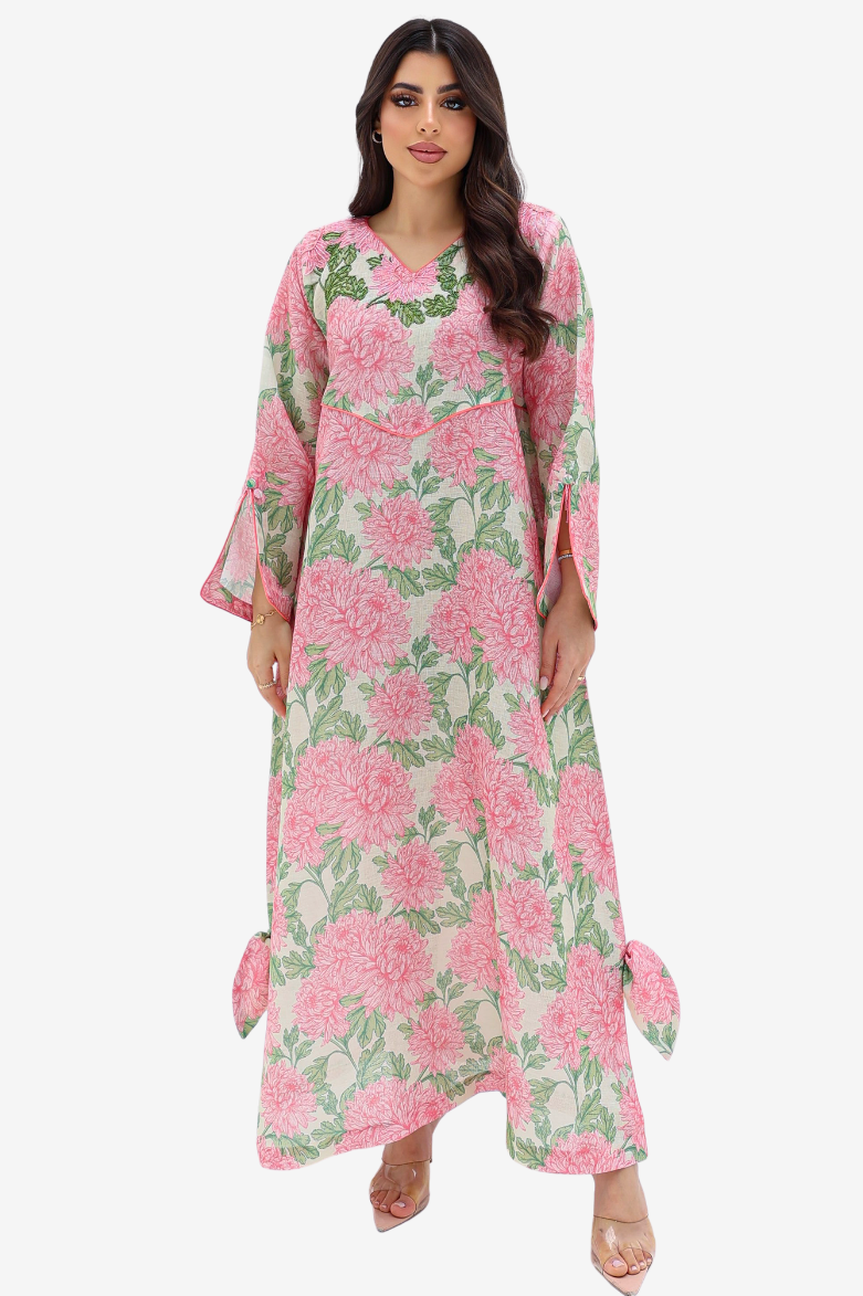 A woman modeling a long-sleeved, floor-length kaftan dress with a pink and green floral print on a light beige background, set against a plain light gray studio backdrop.