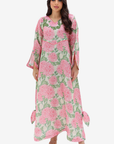 A woman modeling a long-sleeved, floor-length kaftan dress with a pink and green floral print on a light beige background, set against a plain light gray studio backdrop.