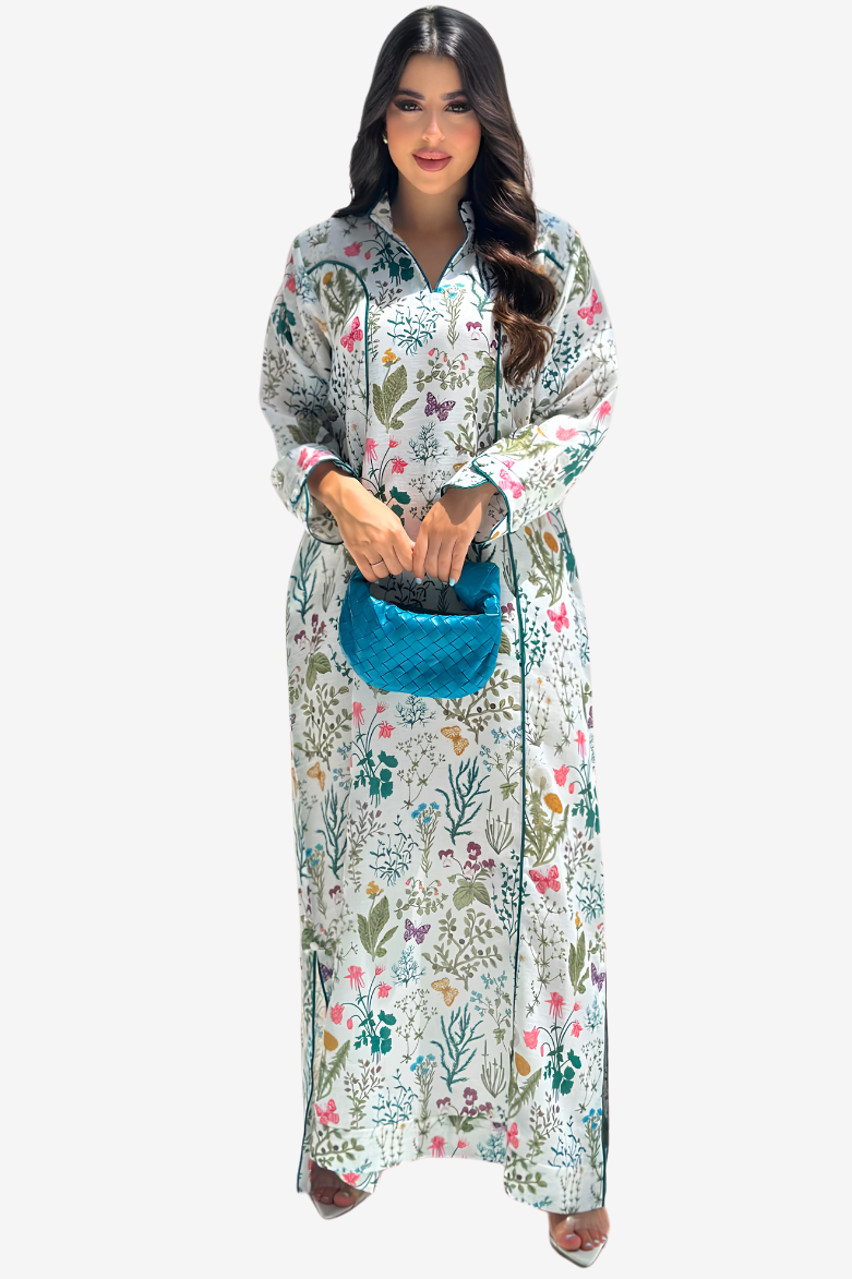 A woman modeling a long-sleeved, button-up maxi dress with a colorful floral and butterfly print on a white background, holding a bright blue textured handbag against a solid light gray background.