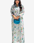 A woman modeling a long-sleeved, button-up maxi dress with a colorful floral and butterfly print on a white background, holding a bright blue textured handbag against a solid light gray background.