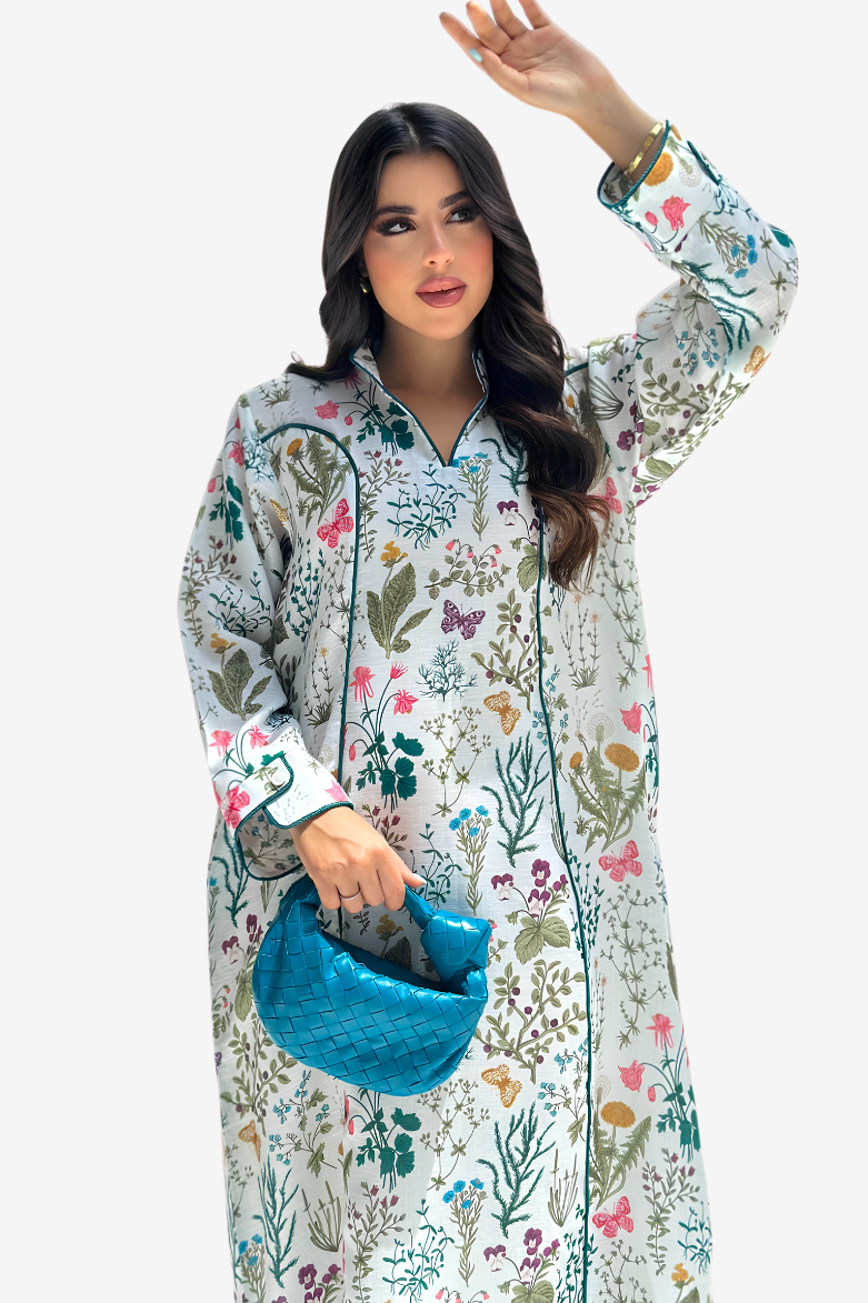 A woman modeling a long-sleeved, button-front kaftan-style nightgown with a colorful botanical and floral print on a white base. She is holding a textured blue clutch bag against a solid light gray background.