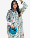 A woman modeling a long-sleeved, button-front kaftan-style nightgown with a colorful botanical and floral print on a white base. She is holding a textured blue clutch bag against a solid light gray background.