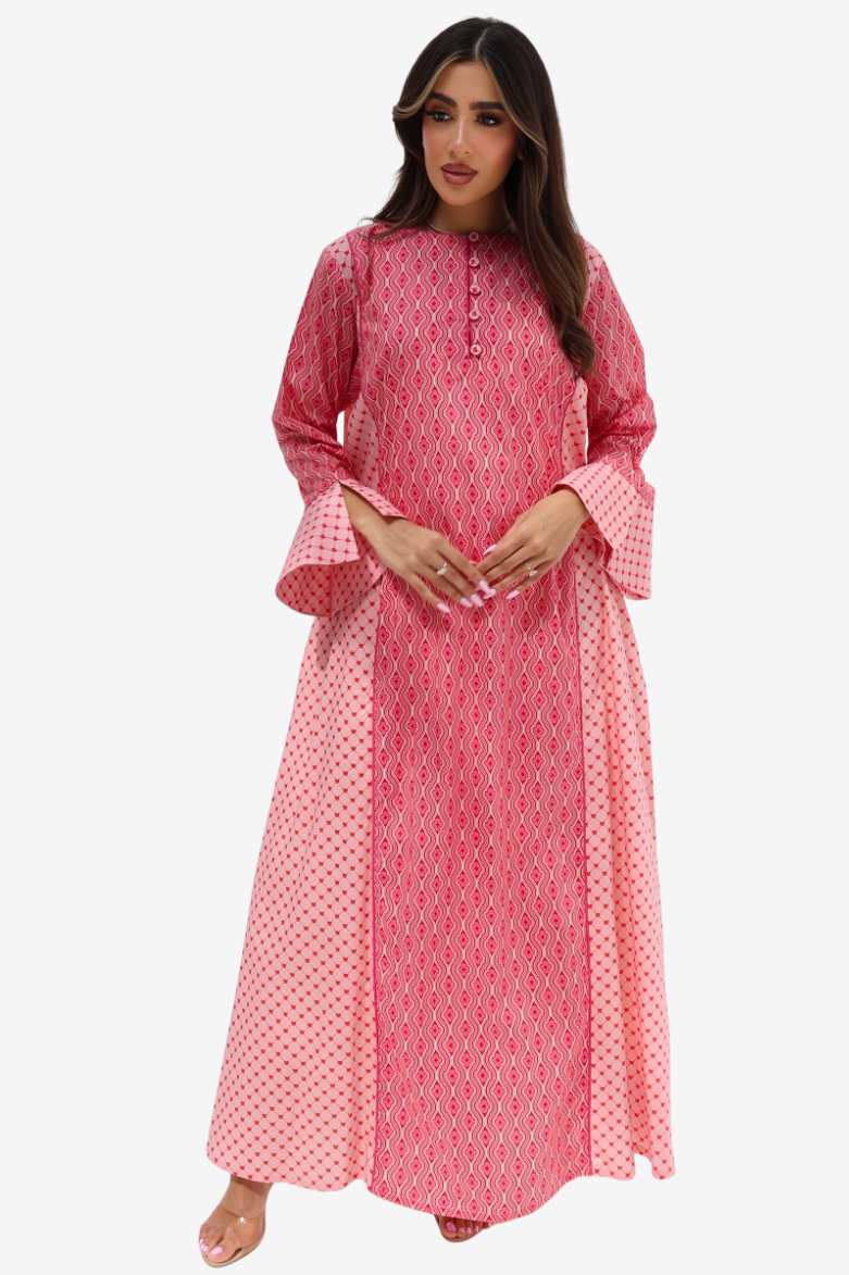 A woman modeling a long pink printed kaftan dress with bell sleeves and a flared hem, photographed against a plain light gray studio background.
