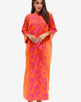 A woman modeling a bright orange and magenta floral print kaftan dress against a plain light gray background.