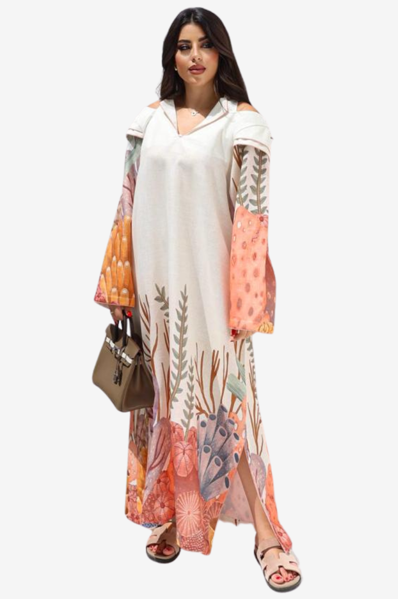 A woman modeling a long white cold-shoulder maxi dress with a colorful botanical and floral print on the sleeves and hem, styled with a brown handbag and sandals against a plain white background.