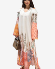 A woman modeling a long white cold-shoulder maxi dress with a colorful botanical and floral print on the sleeves and hem, styled with a brown handbag and sandals against a plain white background.