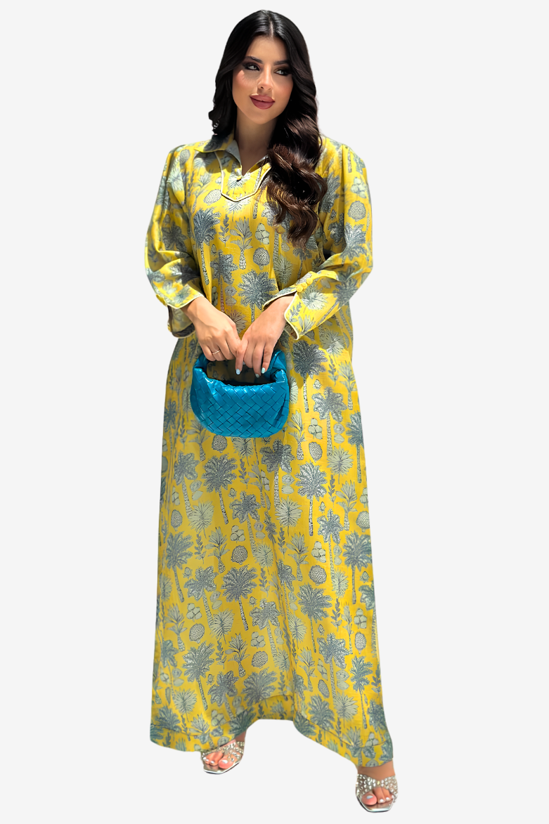 A woman modeling a long yellow kaftan dress with a gray palm tree pattern, holding a small blue handbag against a light gray background.