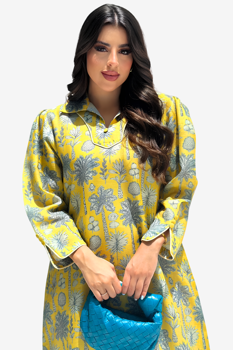 A woman modeling a yellow long-sleeve kurta with a gray botanical pattern, holding a textured blue clutch bag against a light gray background.