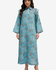 A woman modeling a long-sleeved, floor-length turquoise blue kaftan dress with a beige and gray floral pattern, standing against a light gray background.