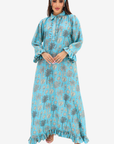 A woman modeling a long-sleeved, high-neck maxi dress in light blue with a gray palm tree and circular pattern, paired with a matching headband, against a plain light gray background.