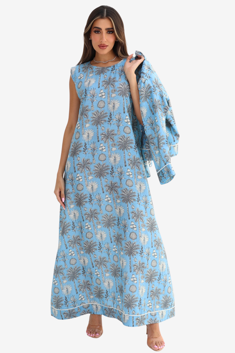 A woman modeling a sleeveless light blue maxi dress with a brown palm tree and circular pattern, featuring a draped shawl over one shoulder, set against a plain light gray background.