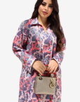 A woman modeling a long-sleeve button-up shirt dress with a purple, pink, and white abstract pattern, holding a small gray handbag against a light gray background.