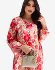 A woman modeling a long-sleeve floral dress with red, pink, and orange flowers on a white background, holding a small gray handbag against a light gray studio backdrop.