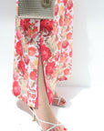 Person wearing a floral dress with a high slit, white sandals, and a silver handbag on a white background.