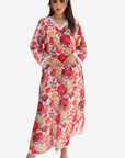 A woman modeling a long-sleeved, V-neck maxi dress with a vibrant red, pink, and orange floral pattern on a white base, paired with a matching floral headband, set against a plain light gray background.