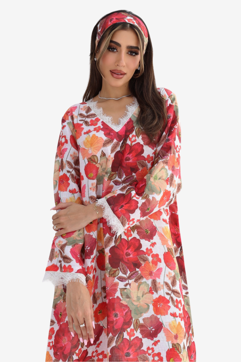 A woman wearing a long-sleeve white kaftan dress with a vibrant red, orange, and green floral print, paired with a matching floral headband, set against a plain light gray background.
