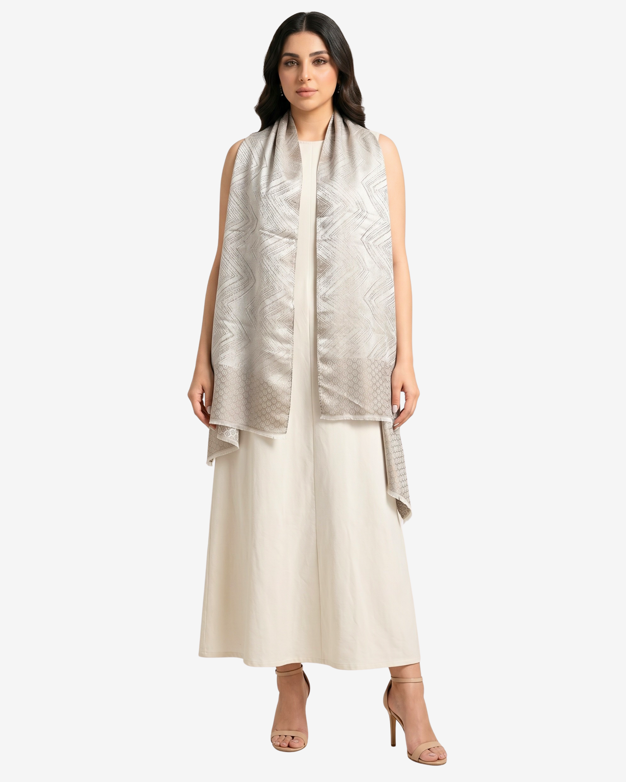 A woman modeling a sleeveless, light beige and silver patterned shawl draped over a cream-colored dress, set against a plain light gray background.