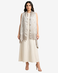 A woman modeling a sleeveless, light beige and silver patterned shawl draped over a cream-colored dress, set against a plain light gray background.