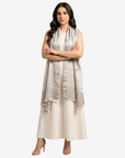 A woman modeling a sleeveless light beige and silver patterned shawl draped over a cream-colored dress, standing against a solid light gray background.