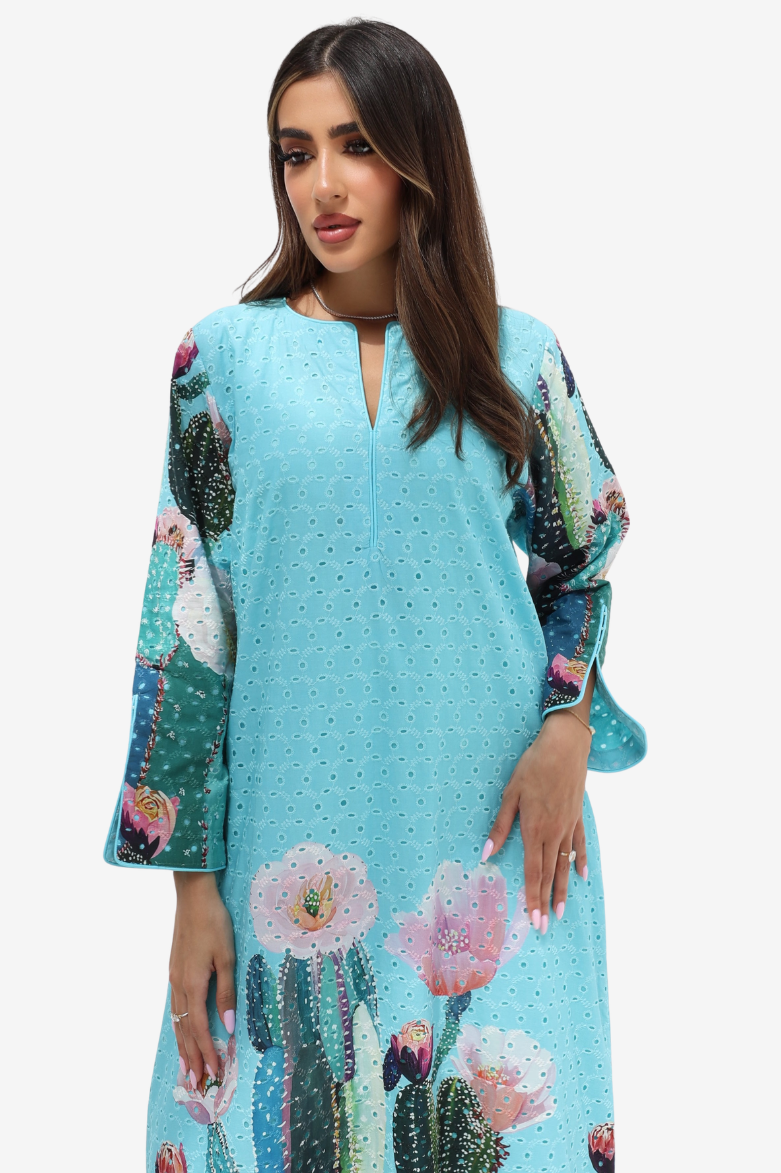 A woman modeling a light blue eyelet kaftan dress with a colorful cactus and flower print on the sleeves and hem, set against a plain white background.