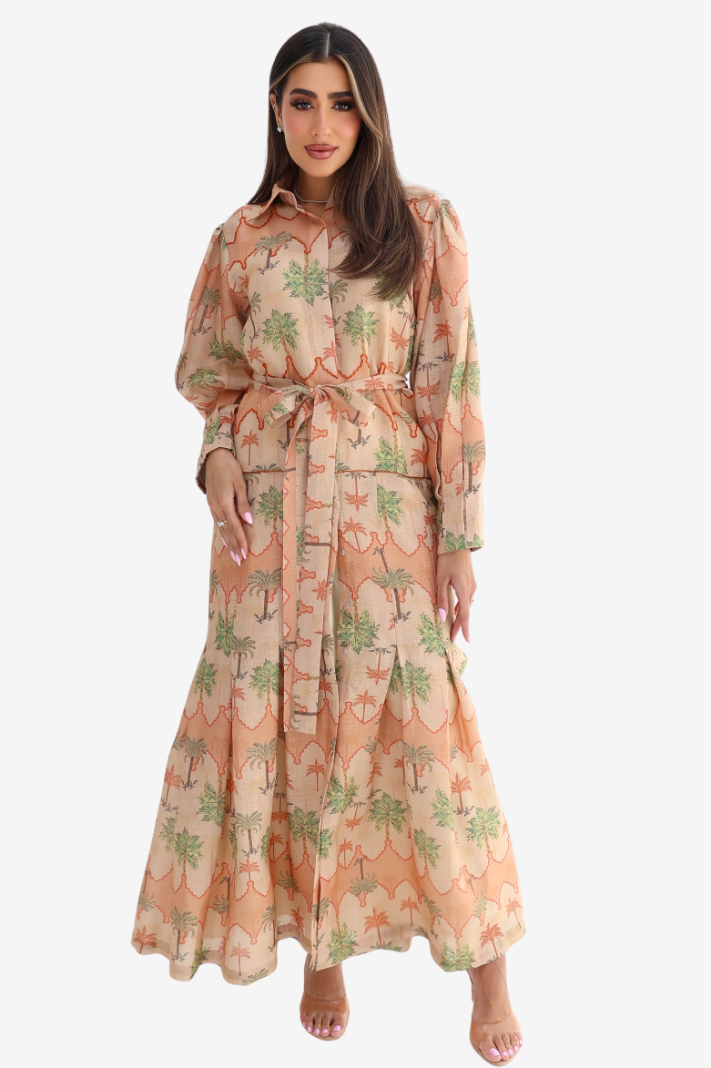 A woman modeling a long-sleeved maxi dress with a beige base and a green palm tree pattern, set against a plain light gray background.