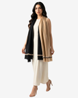 A woman modeling a long beige and black shawl with a geometric patterned border, worn over a cream-colored dress against a light gray background.