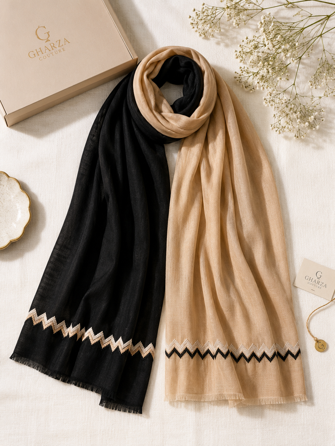 Two lightweight scarves from Gharza London are displayed in a flat-lay arrangement on a textured white fabric background. One scarf is solid black with a beige zigzag border, and the other is solid beige with a black zigzag border.