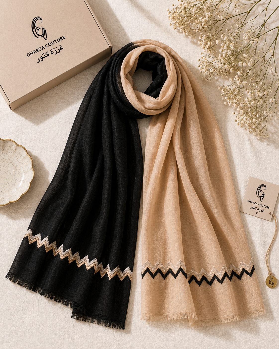 A luxury scarf set from Ghanda Couture, featuring one black scarf and one beige scarf, both with a matching zigzag patterned border. The scarves are styled in a flat-lay arrangement with their branded packaging box, a small decorative dish, and dried baby's breath flowers on a neutral cream background.