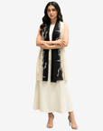 A woman modeling a long cream sleeveless vest over a white dress, accessorized with a black scarf featuring white floral embroidery, against a light gray studio background.