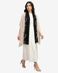 A woman modeling a black scarf with white floral embroidery draped over a white dress, against a light gray studio background.