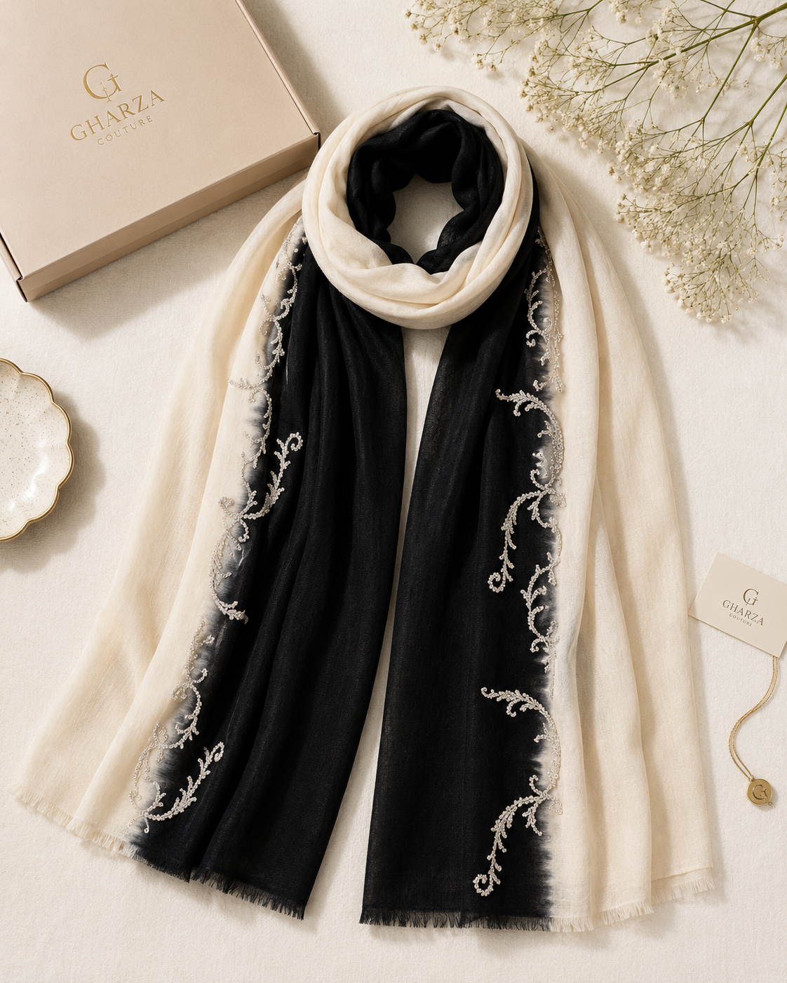 A luxury scarf laid flat on a light beige surface, featuring a black center panel with white embroidered vine motifs and cream-colored fringe and border. A cream-colored Gharza box and a branded hangtag are styled alongside it with dried flowers.