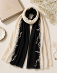 A luxury scarf laid flat on a light beige surface, featuring a black center panel with white embroidered vine motifs and cream-colored fringe and border. A cream-colored Gharza box and a branded hangtag are styled alongside it with dried flowers.