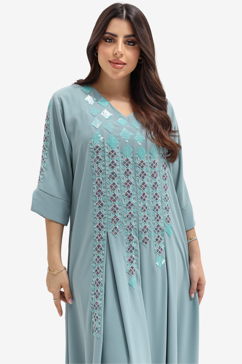 A woman modeling a light blue-green kaftan dress with intricate geometric embroidery in matching and dark purple tones on the chest and sleeves, set against a plain light gray background.