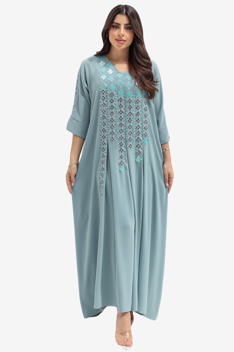 A woman modeling a long, flowing kaftan dress in dusty blue with turquoise and silver geometric embroidery on the chest and down the front, set against a plain light gray background.