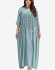 A woman modeling a long, flowing kaftan dress in dusty blue with turquoise and silver geometric embroidery on the chest and down the front, set against a plain light gray background.