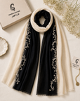 A black and cream scarf with silver floral embroidery, styled on a light beige surface with a branded box, dried flowers, and a small dish.