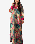 A woman modeling a long-sleeved maxi dress with a dense floral and botanical print in teal, pink, orange, and beige tones, featuring a solid bright pink V-neckline and cuffs.
