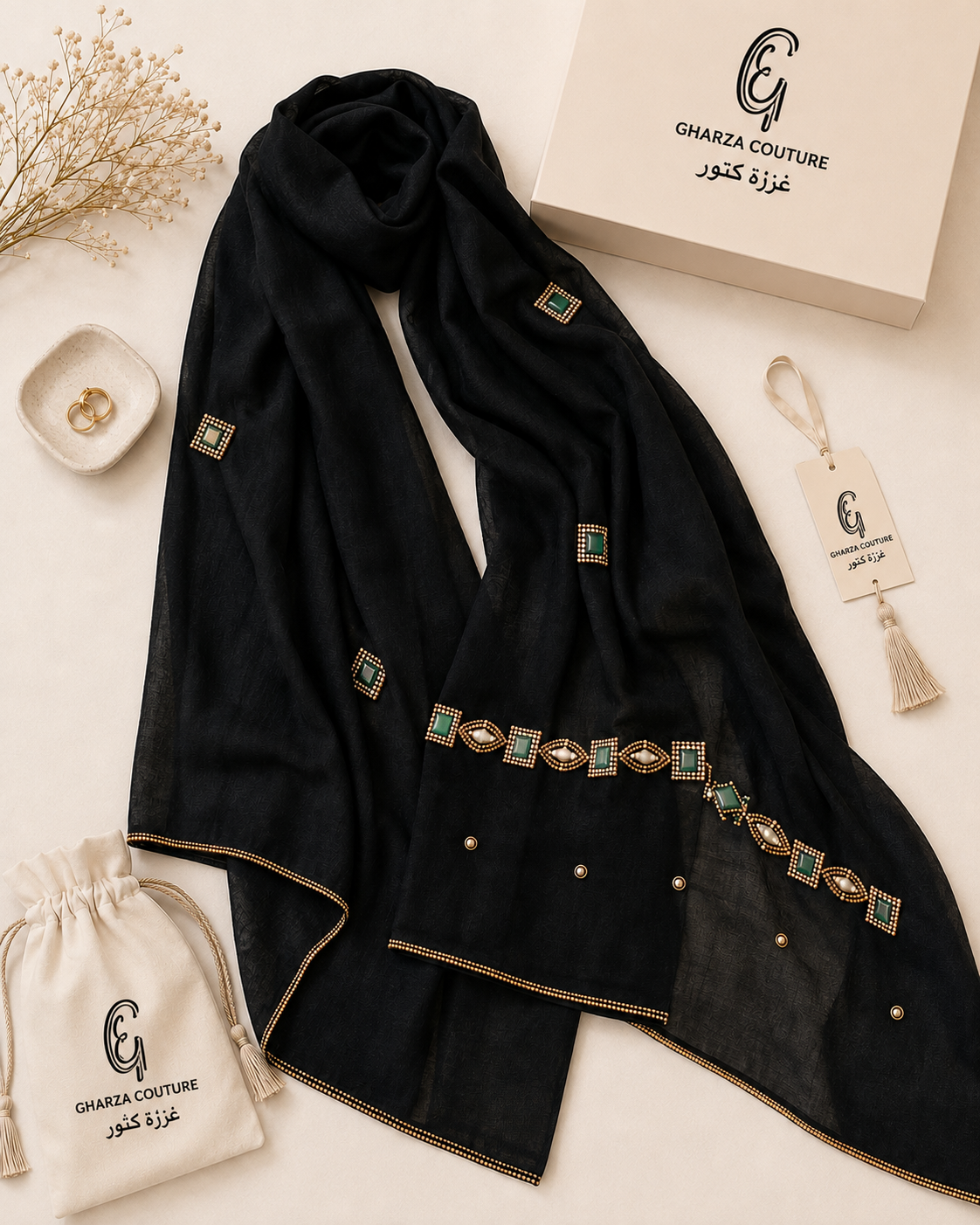 A black sheer scarf with gold trim and green-and-gold embroidered geometric accents, styled in a flat lay with a branded cream box, drawstring pouch, tassel tag, and dried flowers on a light beige background.