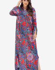 A woman modeling a long-sleeved, floor-length abaya dress with a vibrant red, blue, and black paisley pattern, set against a plain light gray background.