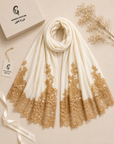 An off-white scarf with intricate gold lace trim at the hem, elegantly draped on a beige background with a branded box, ribbon, and dried floral accents.