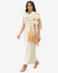 A woman modeling a cream-colored traditional outfit consisting of a long kurta, matching trousers, and a draped dupatta with gold floral lace embroidery, set against a plain light gray background.