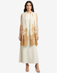 A woman modeling a long cream maxi dress with a matching draped scarf featuring gold floral lace embroidery, set against a plain light gray background.