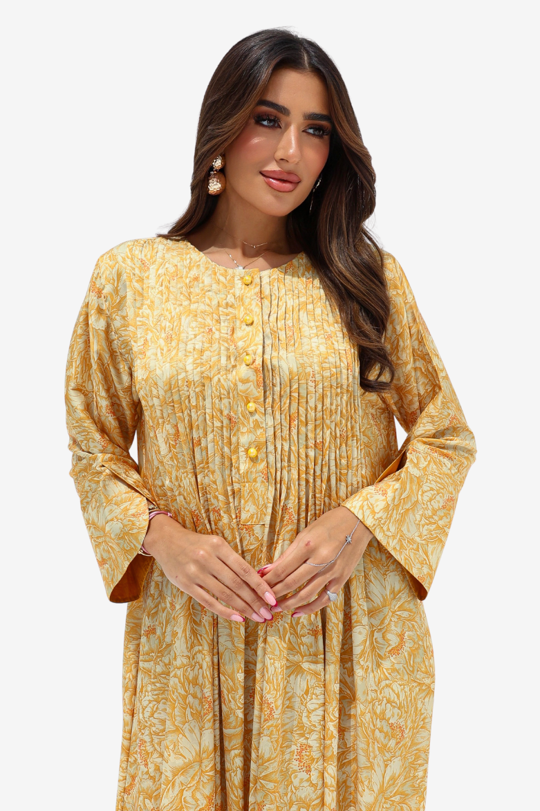 A woman modeling a long-sleeved mustard yellow and beige paisley printed tunic dress with a buttoned front and pleated bodice, set against a plain light gray background.